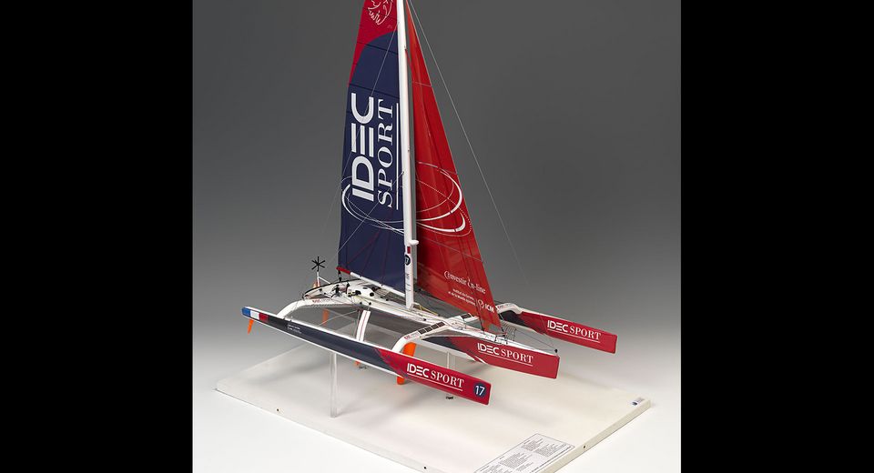 Model sailboat with a red and white sail that has "IDEC SPORT" written on it a white hull, and a red and white striped flag on the top of the mast. The sailboat is mounted on a white base 