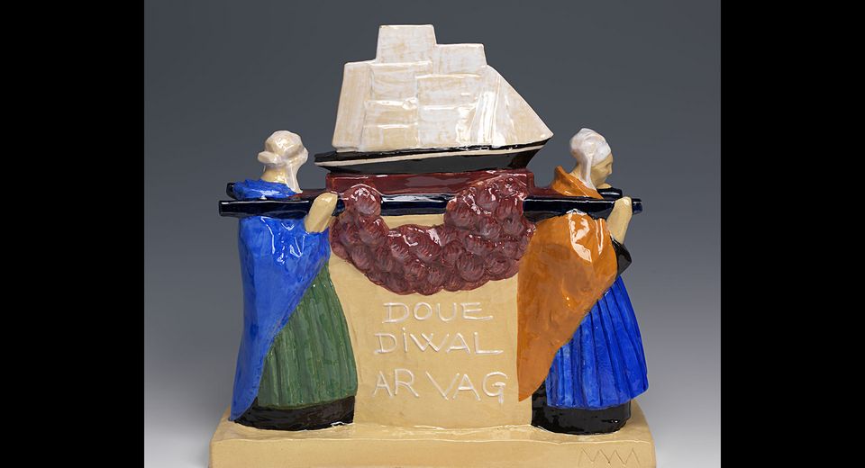 A ceramic sculpture of a ship with a red and purple garland over it. The ship is on a beige base with 'DOUE DIVAL ARAVAG' written in black. The ship is white with a brown mast and sails. There are two figures on either side of the ship. They are wearing blue and green dresses and have white hats. 