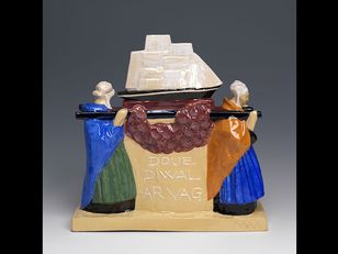 A ceramic sculpture of a ship with a red and purple garland over it. The ship is on a beige base with 'DOUE DIVAL ARAVAG' written in black. The ship is white with a brown mast and sails. There are two figures on either side of the ship. They are wearing blue and green dresses and have white hats. 