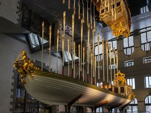 The boat has a figurehead of a golden Neptune. The boat has a golden canopy with a crown on top. The background is a large, open room with high ceilings and windows