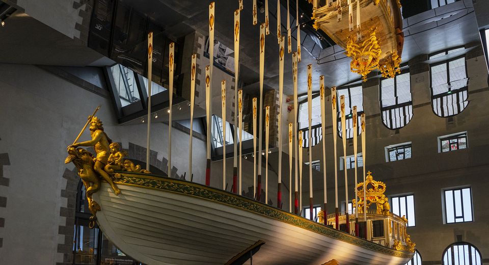 The boat has a figurehead of a golden Neptune. The boat has a golden canopy with a crown on top. The background is a large, open room with high ceilings and windows