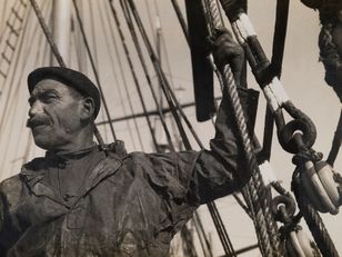  Portrait en contre-plongée d’un marin en ciré et béret, tenant les cordages d’un voilier, entouré de mâts et de poulies – photographie en noir et blanc, début XXe siècle.