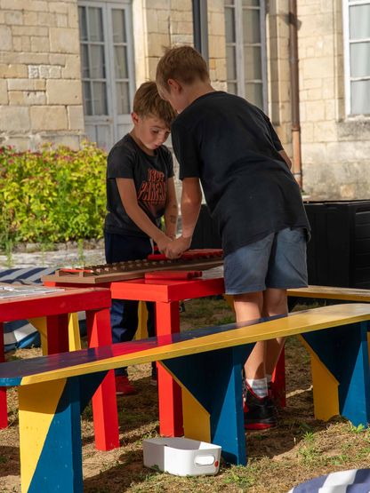 Deux enfants concentrés sur un jeu pédagogique à l’extérieur du musée national de la Marine, manipulant des pièces en bois sur une table rouge.