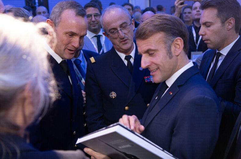 Un officier de marine présente à Emmanuel Macron le livre « Marine(s) » d'Ewan Lebourdais, entouré d'officiers et de personnes assistant à la scène, lors des Assises de l'économie de la mer.