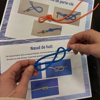 Mains en train de nouer une corde bleue pour apprendre les nœuds marins, avec support pédagogique illustré en arrière-plan
