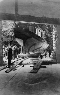 A black and white photograph of a large boat being transported on a wooden ramp. The boat is a wooden vessel with a curved hull and a cabin on top. The ramp is made of wooden planks and is supported by wooden beams. There are several people standing around the boat and ramp, observing the process. The background consists of trees and a stone archway. The photograph appears to be old and has some damage and fading.