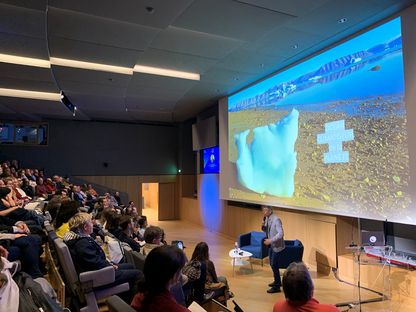 François Sarano s’exprimant devant un amphithéâtre comble au musée national de la Marine, avec la projection d’un iceberg et du texte « Océan climatiseur de la planète » sur grand écran.