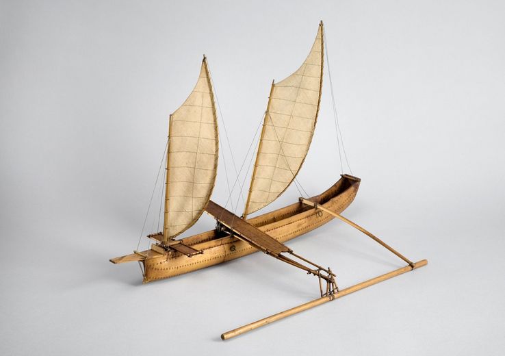 Modèle réduit d’une pirogue à balancier à double voile triangulaire, réalisée en bois clair et toile beige, présentée sur un fond blanc.