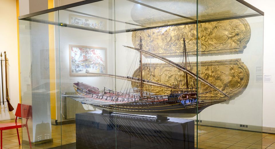 An image of a model ship in a glass display case in the museum, with a white wall and framed artwork in the background