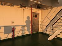 Photographie montrant l’ombre d’une personne projetée sur une paroi à bord d’un navire, avec un escalier métallique visible.