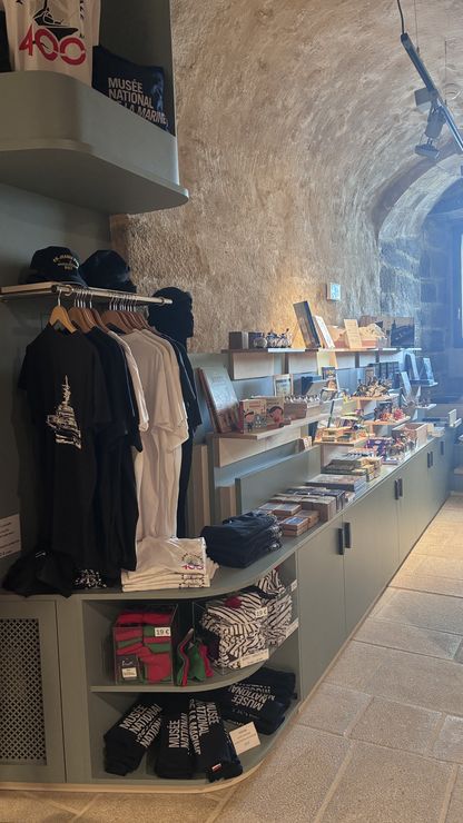 Rayonnage de la boutique du musée national de la Marine de Brest présentant t-shirts, casquettes et vêtements brodés, dans une salle voûtée en pierre.
