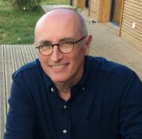 Portrait d’un homme souriant, assis en extérieur sur une terrasse en bois, portant une chemise bleu foncé et des lunettes rondes.