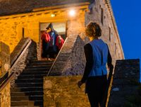Scène nocturne sur un escalier en pierre, silhouettes montant vers un bâtiment fortifié éclairé.