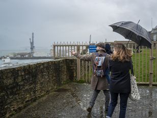 Visiteurs sous la pluie longeant un rempart, l'un d'eux désignant le port et ses grues au loin.