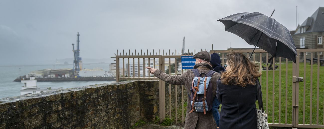 Visiteurs sous la pluie longeant un rempart, l'un d'eux désignant le port et ses grues au loin.