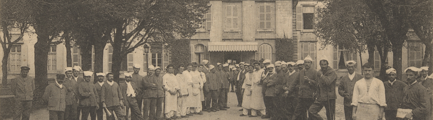 "Carte postale ancienne en noir et blanc montrant l’intérieur de l’Hôpital Maritime de Rochefort-sur-Mer : deux rangées de personnels (militaires en uniforme, infirmiers et aides-soignants en blouse et coiffe blanche) encadrent une allée centrale bordée d’arbres. En arrière-plan, la façade symétrique de l’hôpital, dotée d’un pavillon central à toit mansardé, de lucarnes et d’une horloge sculptée dans le fronton, ainsi qu’un auvent à l’entrée. Deux timbres-poste verts de 5 c. figurent dans les coins supérieurs, et en bas, en lettres rouges, la légende « V.G. 19. ROCHEFORT-sur-MER – Intérieur de l’Hôpital de la Marine »."