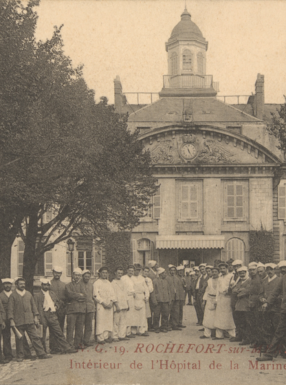 "Carte postale ancienne en noir et blanc montrant l’intérieur de l’Hôpital Maritime de Rochefort-sur-Mer : deux rangées de personnels (militaires en uniforme, infirmiers et aides-soignants en blouse et coiffe blanche) encadrent une allée centrale bordée d’arbres. En arrière-plan, la façade symétrique de l’hôpital, dotée d’un pavillon central à toit mansardé, de lucarnes et d’une horloge sculptée dans le fronton, ainsi qu’un auvent à l’entrée. Deux timbres-poste verts de 5 c. figurent dans les coins supérieurs, et en bas, en lettres rouges, la légende « V.G. 19. ROCHEFORT-sur-MER – Intérieur de l’Hôpital de la Marine »."