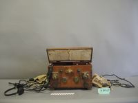 Coffret en bois du téléphone pour scaphandrier fabriqué par Siebe Gorman & Co, posé sur fond blanc. Le boîtier rectangulaire, patiné et usé par le temps, est muni d'une poignée et de sangles de transport en cuir. Un câble électrique enroulé est relié à l'appareil.
