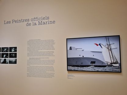 Cimaise de l'exposition « Les Peintres officiels de la Marine » présentant le texte de présentation du corps des POM, une planche-contact de photographies en noir et blanc, et la photographie d'Ewan Lebourdais montrant la frégate Amiral Ronarc'h croisant la goélette Belle Poule.