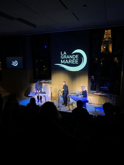 Performance musicale à La Grande Marée au musée national de la Marine : trois musiciens sur scène avec saxophone, percussions et dispositifs électroniques, sous le logo « La Grande Marée », devant un public assis.