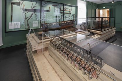 Salle du Dauphin Royal consacrée à la construction navale au Musée national de la Marine à Rochefort, murs verts avec vitrines murales, présentant au premier plan une maquette de coque de navire en construction sur cale et des outils de charpentier de marine exposés.
