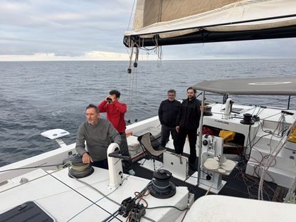 Équipage à bord du catamaran We Explore en navigation au large, sous un ciel nuageux ; un marin à la barre, trois autres observant l’horizon, scène de vie à bord du voilier expérimental de Xavier Veilhan.