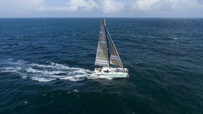 Catamaran de l’expédition We Explore naviguant en pleine mer, vu du ciel, voiles déployées sur l’océan sous un ciel partiellement nuageux.