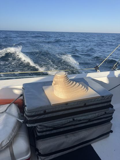 Sculpture lumineuse en forme de coquillage posée sur des coussins sur le pont du voilier We Explore, en mer sous un ciel dégagé.