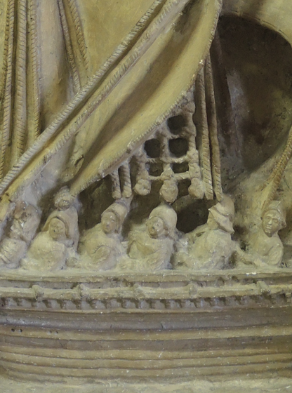 Détail d’une sculpture en pierre montrant la coque d’un bateau ancien, avec des rangées de rames, des personnages et des motifs décoratifs finement sculptés