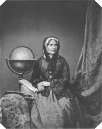Portrait en noir et blanc d’une femme assise près d’un globe terrestre, tenant un livre ou un document sur ses genoux.