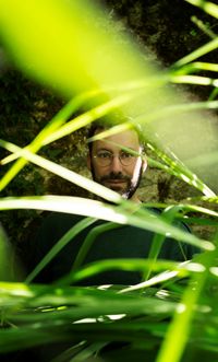 Portrait d’un homme partiellement dissimulé derrière des feuilles vertes, baigné par une lumière naturelle filtrant à travers la végétation.