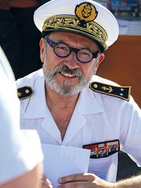 Portrait en gros plan de CA (2S) François Guichard portant une casquette d'officier de marine ornée d'insignes dorés, en discussion avec une personne au premier plan.
