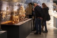 Visiteurs observant une vitrine d'objets et maquettes dorées, dans une salle d'exposition aux murs illustrés.
