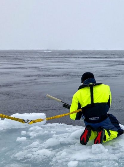 Océanographe en combinaison de sécurité effectuant des relevés sur la banquise arctique, face à un navire dans le brouillard, en mer de Kara.