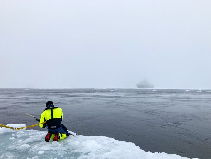 Océanographe en combinaison de sécurité effectuant des relevés sur la banquise arctique, face à un navire dans le brouillard, en mer de Kara.