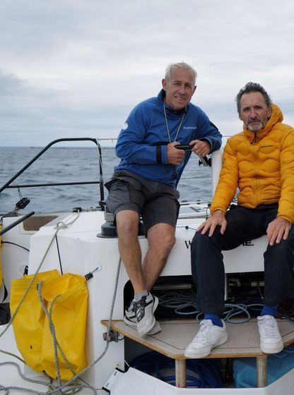 Xavier Veilhan et Roland Jourdain à l’arrière du catamaran We Explore, en mer, vêtus de tenues techniques, regardant l’objectif.