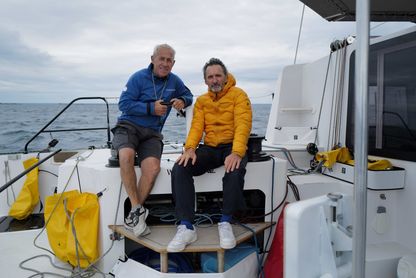 Xavier Veilhan et Roland Jourdain à l’arrière du catamaran We Explore, en mer, vêtus de tenues techniques, regardant l’objectif.