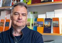 Portrait de Marc Wiltz, fondateur des éditions Magellan & Cie, devant une étagère remplie de livres de sa maison d’édition.