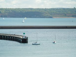 Rade de Brest avec jetée en pierre, voilier et embarcation sur l'eau, côte verdoyante à l'horizon.