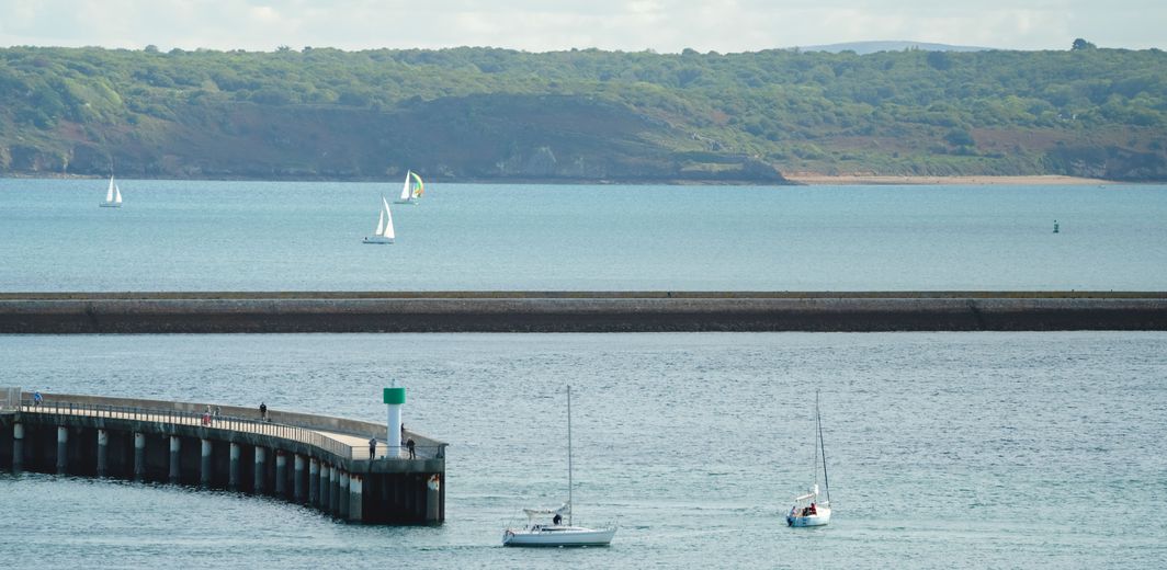 Rade de Brest avec jetée en pierre, voilier et embarcation sur l'eau, côte verdoyante à l'horizon.