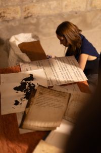 Documents anciens, feuillets manuscrits et carte du monde posés sur une table en bois, avec en arrière-plan une jeune femme concentrée, assise au sol dans une pièce aux murs en pierre, ambiance tamisée évoquant une exploration ou un travail de recherche historique.