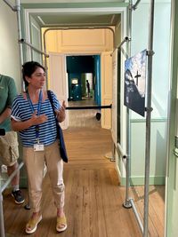 L'administratrice du musée national de la Marine à Rochefort et commissaire de l'exposition, sourit dans un couloir aux murs clairs et au parquet en bois. Elle porte un haut bleu à motifs, un pantalon clair et un sac en bandoulière. Des dispositifs de présentation de l'exposition sont visibles derrière elle.