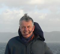 Portrait d’un homme souriant sur un bateau, portant une veste sombre, avec une mer agitée et une île montagneuse en arrière-plan.