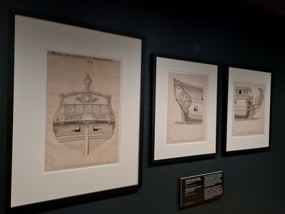 Trois dessins encadrés de style ornemental, mêlant figures humaines et motifs décoratifs, présentés sur un mur sombre d’exposition