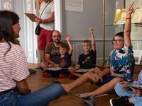 Groupe d’enfants assis sur un tapis participant à une visite-jeu, levant la main face à une médiatrice dans une salle du musée
