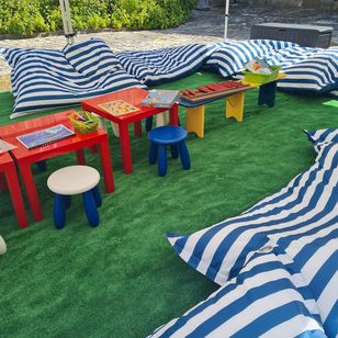 Espace ludique en plein air avec tables de jeux colorées, coussins rayés bleu et blanc et matériel pédagogique à destination des enfants.