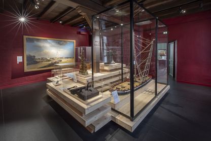 Salle des machines du parcours permanent rénové du Musée national de la Marine à Rochefort, murs rouge foncé, avec une peinture marine encadrée au mur, une vitrine haute abritant un mât ou dispositif technique et une vitrine centrale présentant des maquettes et objets liés à la propulsion navale.