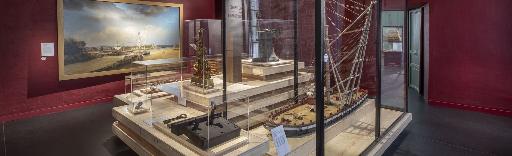 Salle des machines du parcours permanent rénové du Musée national de la Marine à Rochefort, murs rouge foncé, avec une peinture marine encadrée au mur, une vitrine haute abritant un mât ou dispositif technique et une vitrine centrale présentant des maquettes et objets liés à la propulsion navale.