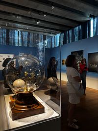 Planétaire mécanique du XIXe siècle présenté sous cloche de verre dans une vitrine d’exposition, au sein d’une salle sombre aux murs bleus, avec visiteurs observant les œuvres en arrière-plan.