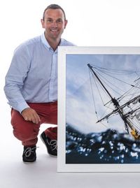 Ewan Lebourdais, photographe et Peintre officiel de la Marine, pose agenouillé à côté d'une photographie encadrée représentant le mât et le gréement d'un voilier au-dessus de l'eau.